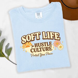 May include: Light blue t-shirt with the words "SOFT LIFE" above "HUSTLE CULTURE" and "Protect Your Peace" in a retro design. The shirt also features a sun and flower graphic.