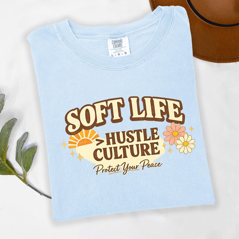 May include: Light blue t-shirt with the words "SOFT LIFE" above "HUSTLE CULTURE" and "Protect Your Peace" in a retro design. The shirt also features a sun and flower graphic.