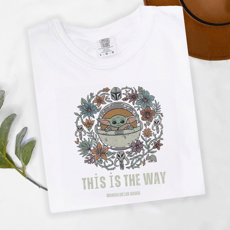 May include: White t-shirt featuring a graphic of Grogu from The Mandalorian series. The design includes Grogu in a pod surrounded by flowers and the text "THIS IS THE WAY". The shirt is labeled "Comfort Colors".