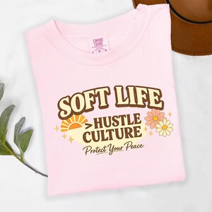 May include: A light pink t-shirt with the words "SOFT LIFE" in a brown retro font. Below it, the text reads "HUSTLE CULTURE" and "Protect Your Peace". The shirt also features a sun graphic and floral accents.