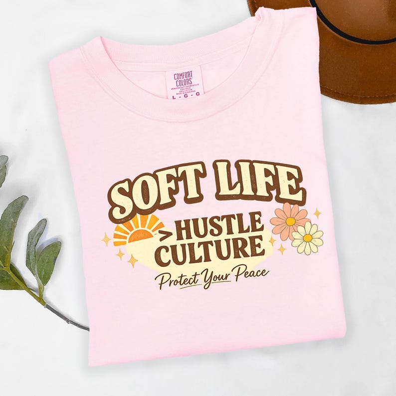 May include: A light pink t-shirt with the words "SOFT LIFE" in a brown retro font. Below it, the text reads "HUSTLE CULTURE" and "Protect Your Peace". The shirt also features a sun graphic and floral accents.