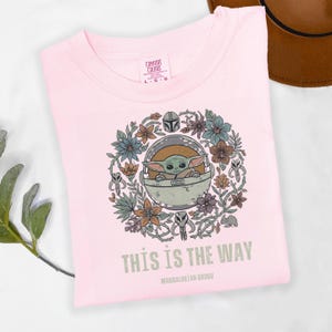 May include: Light pink t-shirt featuring a graphic of Grogu in a pod, surrounded by flowers and Mandalorian helmet. The text "THIS IS THE WAY" and "MANDALORIAN GROGU" are printed below the graphic. The shirt is labeled "Comfort Colors".