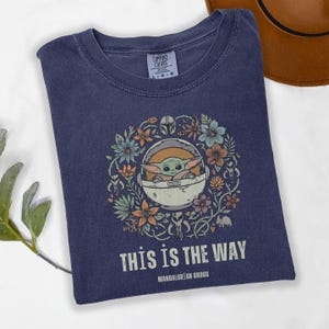 May include: Navy blue t-shirt featuring a graphic of Grogu in a floral wreath, with the text "THIS IS THE WAY" and "MANDALORIAN GROGU". The shirt is a Comfort Colors brand.