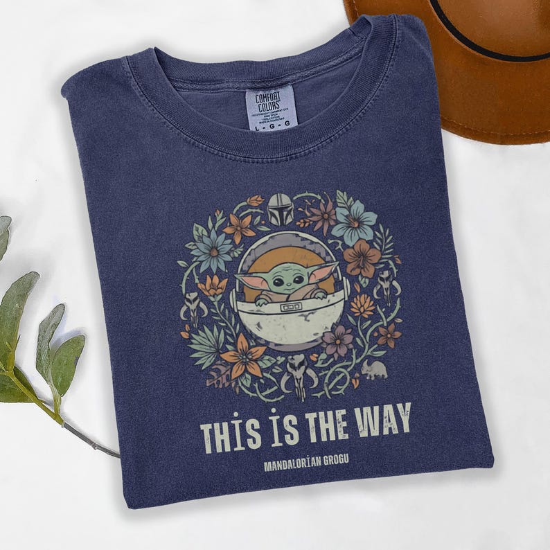 May include: Navy blue t-shirt featuring a graphic of Grogu in a floral wreath, with the text "THIS IS THE WAY" and "MANDALORIAN GROGU". The shirt is a Comfort Colors brand.