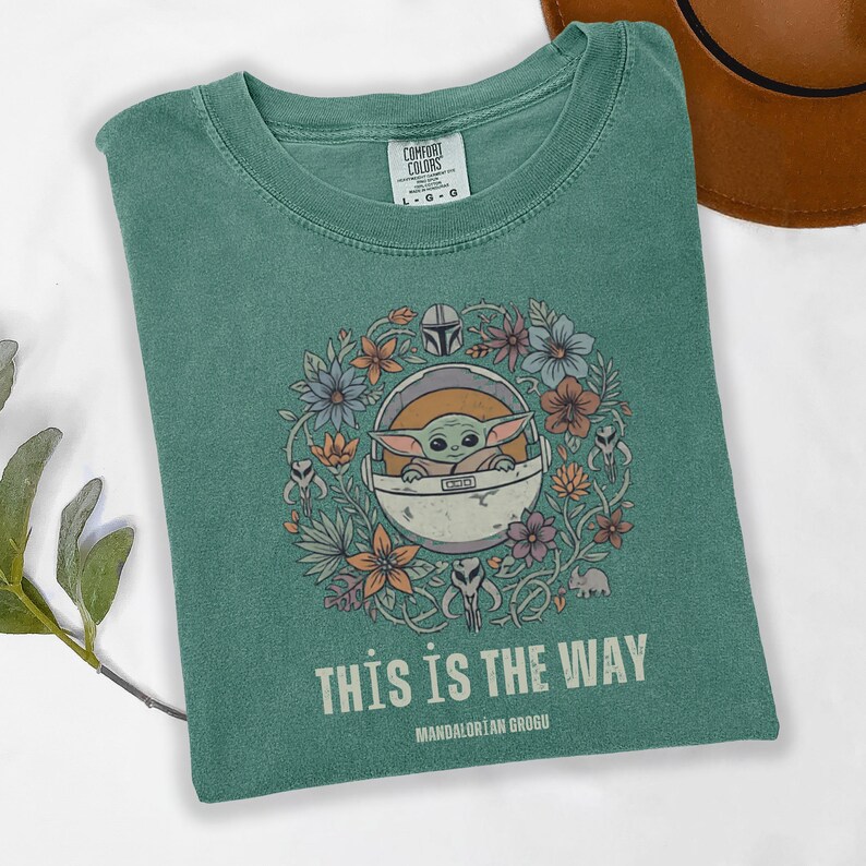 May include: Teal green t-shirt featuring a graphic of Grogu in a pod surrounded by flowers. The text "THIS IS THE WAY" and "MANDALORIAN GROGU" are printed below the graphic. The shirt is a Comfort Colors brand.