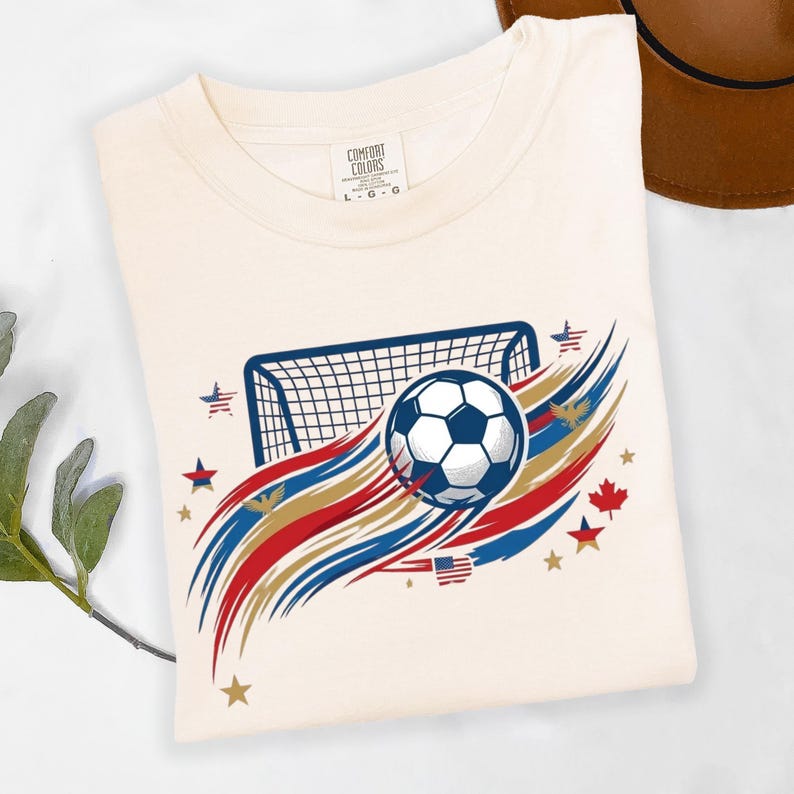 May include: A cream-colored short-sleeve t-shirt featuring a graphic of a soccer ball in front of a goal net. The design includes red, white, and blue brushstroke accents, small stars, a maple leaf, and a small American flag icon on the front.