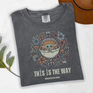 May include: Gray t-shirt featuring a floral design with Grogu in a pod, surrounded by flowers in shades of blue, orange, and purple. The text "THIS IS THE WAY" and "MANDALORIAN GROGU" are printed below the image.