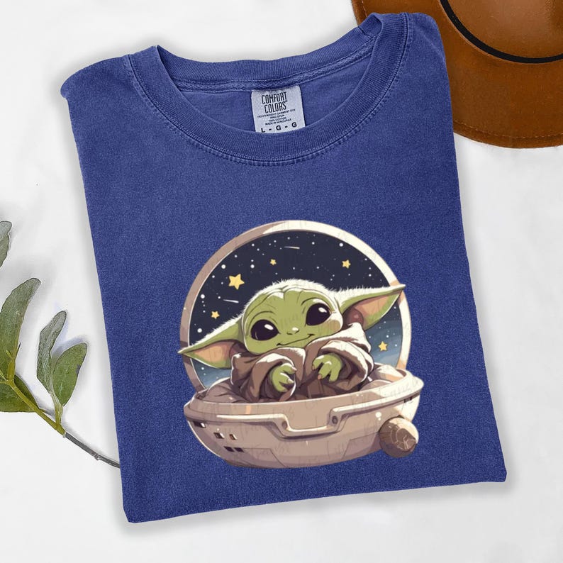 Puede incluir: Camiseta azul con un gr&aacute;fico de un personaje verde alien&iacute;gena en una nave espacial sobre un fondo estrellado. La camiseta tiene cuello redondo y mangas cortas. La etiqueta de la marca "Comfort Colors" es visible.