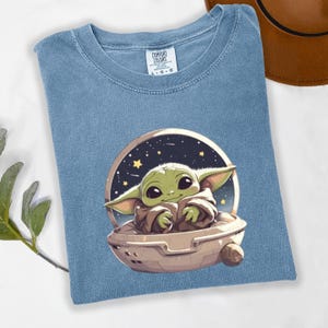 Puede incluir: Camiseta azul claro con un gr&aacute;fico de un personaje verde alien&iacute;gena en una c&aacute;psula espacial sobre un fondo estrellado. La camiseta tiene cuello redondo y mangas cortas. La etiqueta dice "COMFORT COLORS" y las tallas son L-G-G.