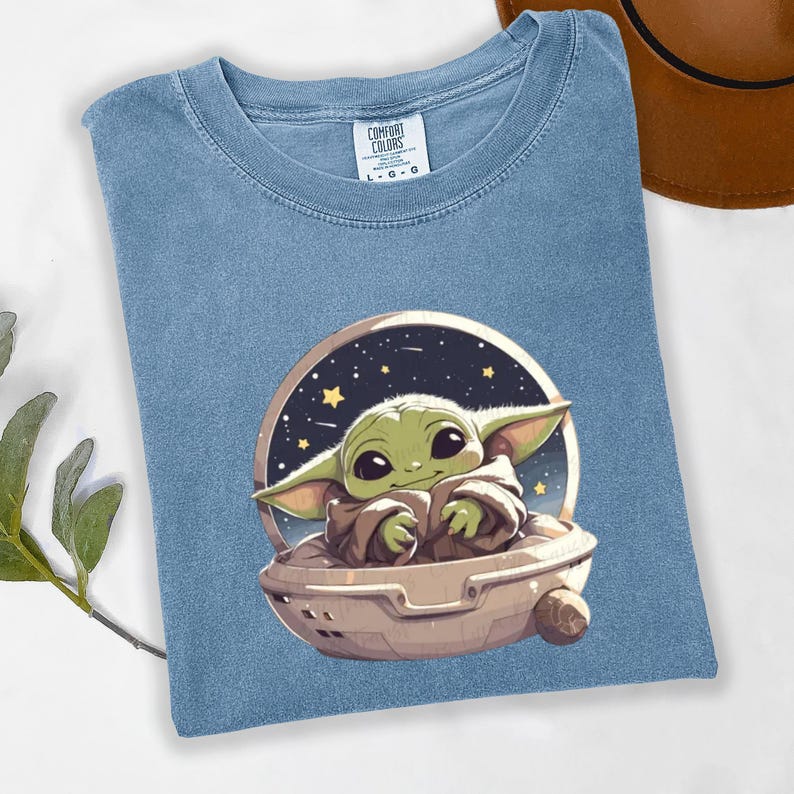 Puede incluir: Camiseta azul claro con un gr&aacute;fico de un personaje verde alien&iacute;gena en una c&aacute;psula espacial sobre un fondo estrellado. La camiseta tiene cuello redondo y mangas cortas. La etiqueta dice "COMFORT COLORS" y las tallas son L-G-G.