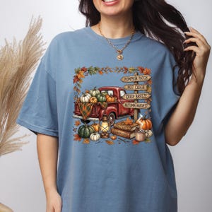 Puede incluir: Una camiseta azul polvoriento con un gr&aacute;fico de una camioneta roja llena de calabazas, una linterna y libros. Un poste de madera muestra el texto: PUMPKIN PATCH, HOT CIDER, CRISP BREEZE, AUTUMN HARVEST. El dise&ntilde;o est&aacute; enmarcado por hojas de oto&ntilde;o.
