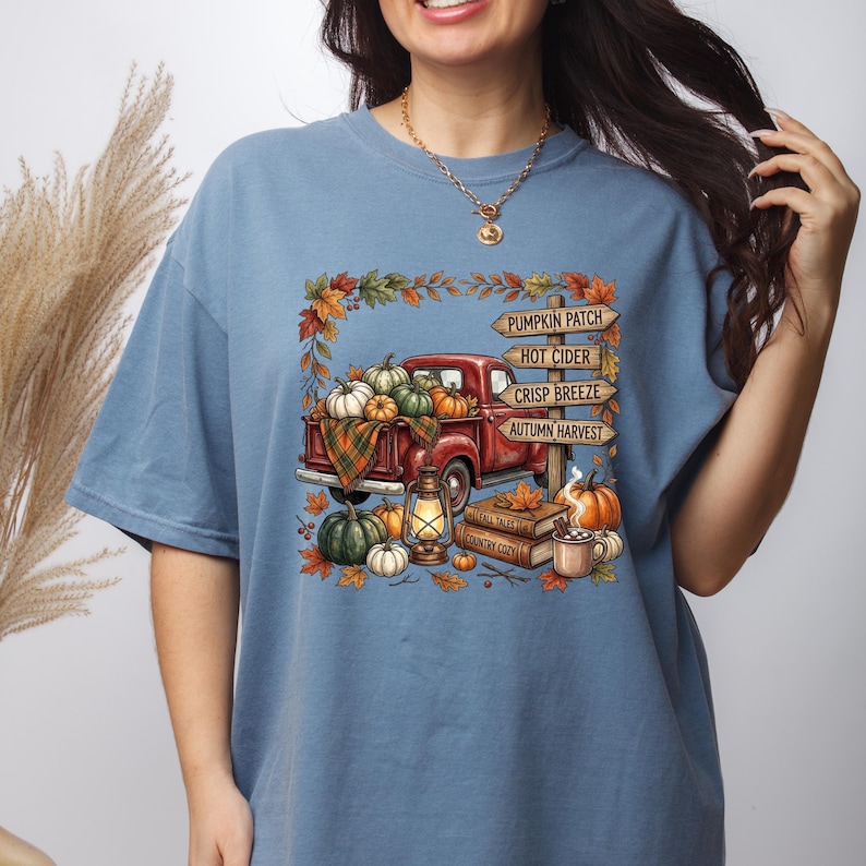 Puede incluir: Una camiseta azul polvoriento con un gr&aacute;fico de una camioneta roja llena de calabazas, una linterna y libros. Un poste de madera muestra el texto: PUMPKIN PATCH, HOT CIDER, CRISP BREEZE, AUTUMN HARVEST. El dise&ntilde;o est&aacute; enmarcado por hojas de oto&ntilde;o.