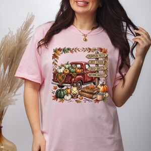 Puede incluir: Una camiseta rosa claro con un gr&aacute;fico de una camioneta roja cargada de calabazas, una linterna y libros. Un poste de madera muestra el texto Pumpkin Patch, Hot Cider, Crisp Breeze y Autumn Harvest, rodeado de hojas de oto&ntilde;o.