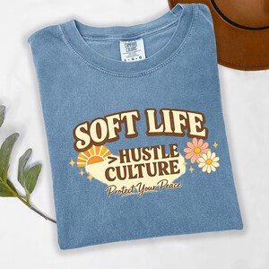 May include: A blue t-shirt with the words "SOFT LIFE HUSTLE CULTURE Protect Your Peace" in a retro design. The graphic includes a sun, flowers, and stars. The shirt is folded.