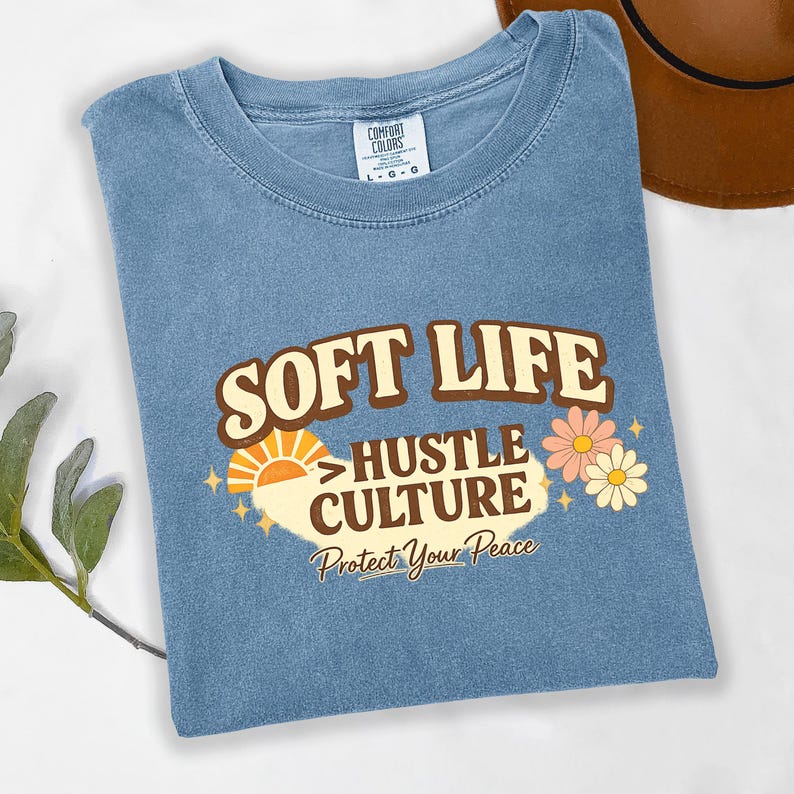 May include: A blue t-shirt with the words "SOFT LIFE HUSTLE CULTURE Protect Your Peace" in a retro design. The graphic includes a sun, flowers, and stars. The shirt is folded.