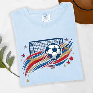 May include: A light blue t-shirt featuring a graphic of a soccer ball and a goal net. The design includes red, white, and blue brushstroke accents, small stars, and various national flag icons. The inner neck label displays the text Comfort Colors L-G-G.