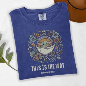 May include: A blue t-shirt featuring a graphic of Grogu in a pod surrounded by flowers. The text "THIS IS THE WAY" and "MANDALORIAN GROGU" are printed below the graphic. The shirt is a Comfort Colors brand, size L-G.