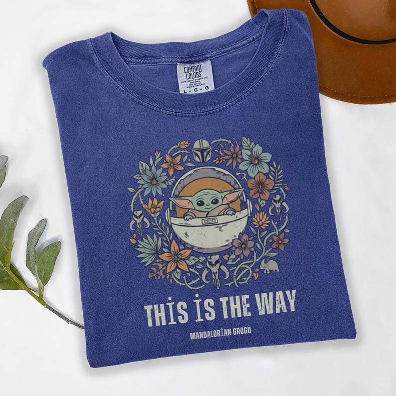 May include: A blue t-shirt featuring a graphic of Grogu in a pod surrounded by flowers. The text "THIS IS THE WAY" and "MANDALORIAN GROGU" are printed below the graphic. The shirt is a Comfort Colors brand, size L-G.
