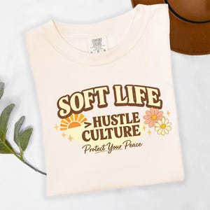 May include: Cream-colored t-shirt with the words "SOFT LIFE" in brown retro font. Below, it reads "HUSTLE CULTURE" and "Protect Your Peace". The design includes a sun graphic and flowers.