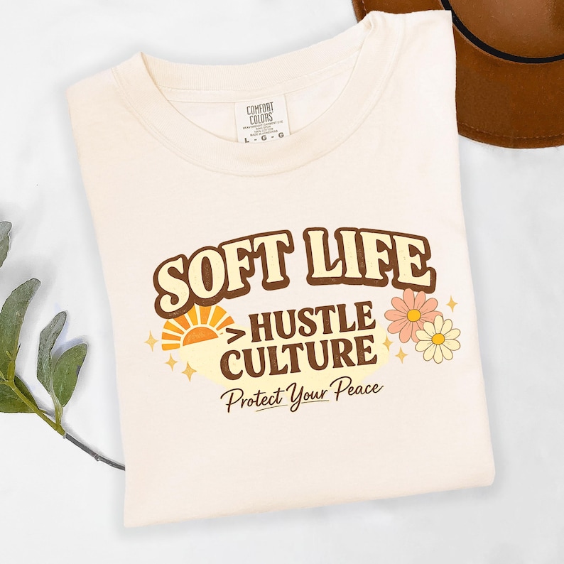 May include: Cream-colored t-shirt with the words "SOFT LIFE" in brown retro font. Below, it reads "HUSTLE CULTURE" and "Protect Your Peace". The design includes a sun graphic and flowers.