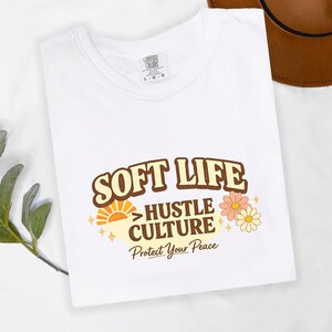 May include: White t-shirt with the words "SOFT LIFE HUSTLE CULTURE Protect Your Peace" in a retro font. The design includes a sun, stars, and flowers in shades of orange, yellow, and pink. The shirt is folded.