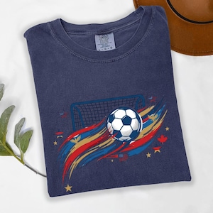 May include: A navy blue t-shirt featuring a graphic of a soccer ball in front of a goal net, surrounded by red, gold, and blue brushstroke accents, small stars, and maple leaf icons. The inner neck label displays the text Comfort Colors L-G-G.