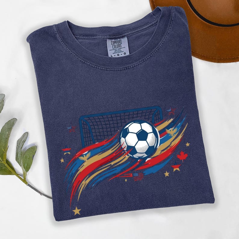 May include: A navy blue t-shirt featuring a graphic of a soccer ball in front of a goal net, surrounded by red, gold, and blue brushstroke accents, small stars, and maple leaf icons. The inner neck label displays the text Comfort Colors L-G-G.