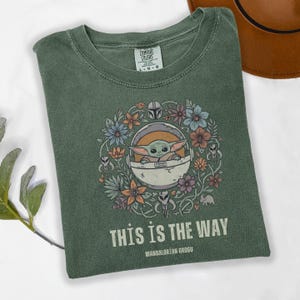May include: Green t-shirt with a floral design surrounding a Grogu character in a pod. The text "THIS IS THE WAY" and "MANDALORIAN GROGU" are printed below the graphic. The shirt is made by Comfort Colors.