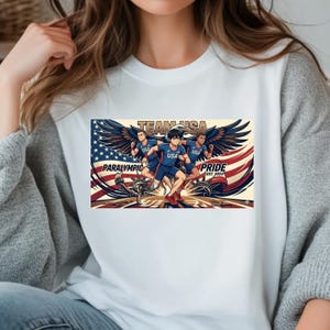 May include: White t-shirt featuring a graphic of Team USA Paralympians with angel wings, running in front of an American flag design. The text includes "Team USA", "Paralympic", and "Pride Est. 2024". The design is in shades of blue, red, and gold.