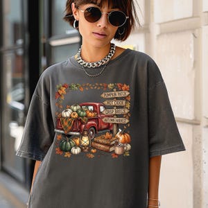 Puede incluir: Una camiseta gris carb&oacute;n con un gr&aacute;fico de una camioneta roja llena de calabazas, una linterna, libros apilados y una taza. Un poste de madera muestra el texto: PUMPKIN PATCH, HOT CIDER, CRISP BREEZE, AUTUMN HARVEST, FALL TALES, COUNTRY COZY.