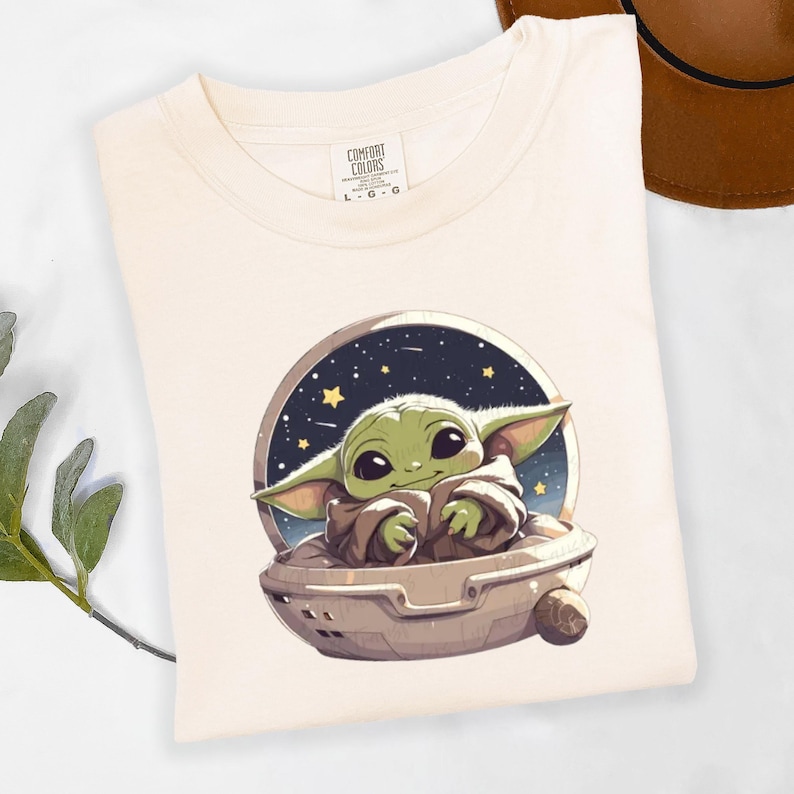Puede incluir: Camiseta color crema con una imagen de dibujos animados de un personaje verde parecido a un extraterrestre en una nave espacial sobre un fondo estrellado. La camiseta tiene una etiqueta Comfort Colors y est&aacute; doblada. Un sombrero marr&oacute;n es visible en el fondo.
