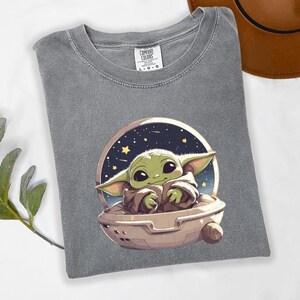 Puede incluir: Camiseta gris Comfort Colors con una imagen de dibujos animados de un personaje alien&iacute;gena verde en una nave espacial sobre un fondo estrellado. La camiseta est&aacute; doblada y tiene una etiqueta en el centro superior.