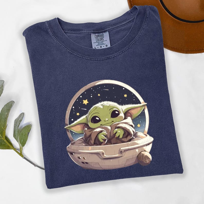 Puede incluir: Camiseta azul marino con una ilustraci&oacute;n de dibujos animados de un personaje alien&iacute;gena verde en una c&aacute;psula contra un fondo estrellado. La camiseta est&aacute; hecha de un material suave y tiene la etiqueta de la marca Comfort Colors.