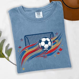 May include: A blue short-sleeve t-shirt with a graphic print featuring a soccer ball, a goal net, and red, gold, and blue brushstroke accents. The design includes small stars, eagles, and flags. The neck label reads Comfort Colors L G G.