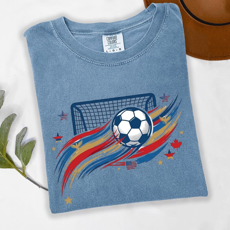 May include: A blue short-sleeve t-shirt with a graphic print featuring a soccer ball, a goal net, and red, gold, and blue brushstroke accents. The design includes small stars, eagles, and flags. The neck label reads Comfort Colors L G G.
