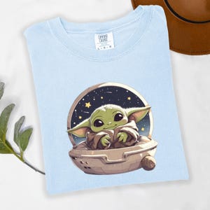 Puede incluir: Camiseta azul claro con un gr&aacute;fico de un personaje alien&iacute;gena verde en una c&aacute;psula espacial sobre un fondo estrellado. La camiseta tiene cuello redondo y mangas cortas. La marca "Comfort Colors" es visible.