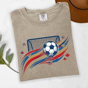 May include: A beige short-sleeve t-shirt featuring a graphic of a soccer ball, a goal net, and abstract red, white, and blue brushstrokes. The design includes small icons of stars, eagles, and a maple leaf. The neck label reads Comfort Colors L G G.