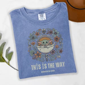 May include: A blue t-shirt featuring a graphic of Grogu, surrounded by flowers, in a pod. The text "THIS IS THE WAY" and "MANDALORIAN GROGU" are printed below the graphic. The shirt has a crew neck and short sleeves.