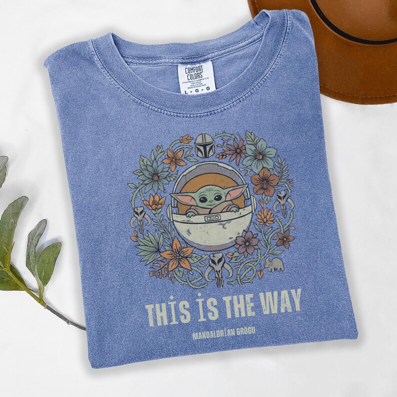May include: A blue t-shirt featuring a graphic of Grogu, surrounded by flowers, in a pod. The text "THIS IS THE WAY" and "MANDALORIAN GROGU" are printed below the graphic. The shirt has a crew neck and short sleeves.