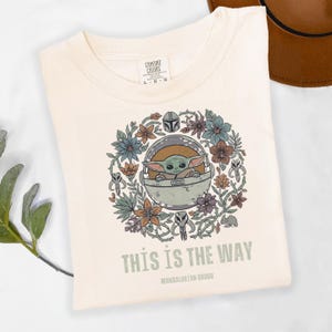 May include: Cream-colored t-shirt featuring a circular design with Grogu in a pod, surrounded by flowers and Mandalorian symbols. The text "THIS IS THE WAY" and "MANDALORIAN GROGU" are printed below the design. The shirt is folded.