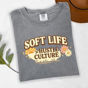 May include: A gray Comfort Colors t-shirt with the words "SOFT LIFE" above "HUSTLE CULTURE" and "Protect Your Peace" in a retro font. The design includes a sun, flowers, and stars.