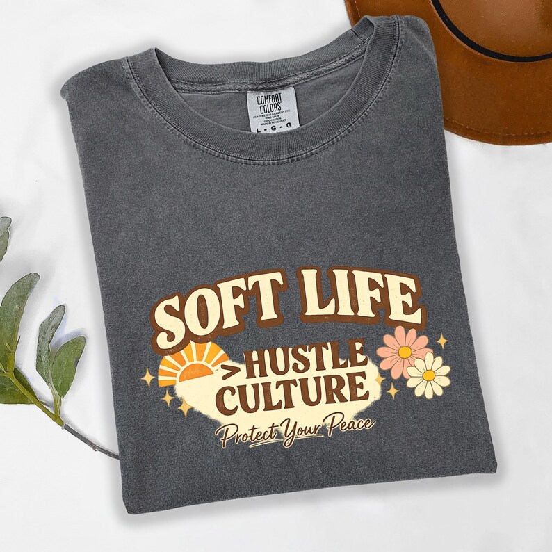 May include: Dark gray Comfort Colors t-shirt with the words "SOFT LIFE" and "HUSTLE CULTURE" in a retro font. The design includes a sun, flowers, and the phrase "Protect Your Peace".