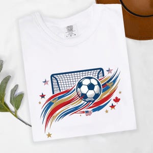 May include: A white t-shirt featuring a graphic of a soccer ball and a goal net. The design includes red, white, and blue brushstroke accents, small stars, and eagle silhouettes. The inner neck label displays the text Comfort Colors L G G.
