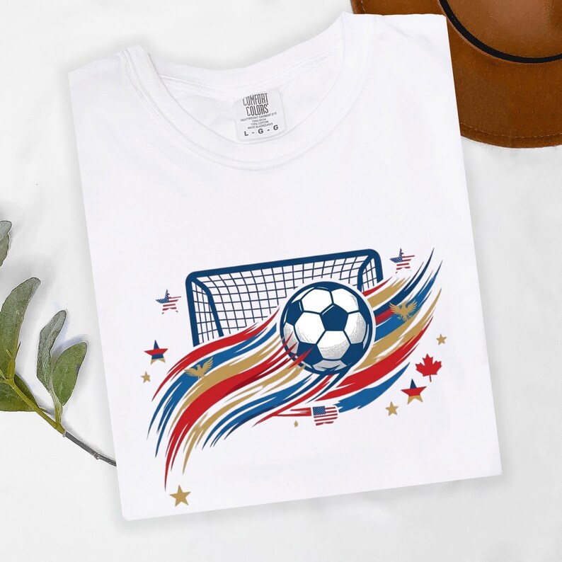 May include: A white t-shirt featuring a graphic of a soccer ball and a goal net. The design includes red, white, and blue brushstroke accents, small stars, and eagle silhouettes. The inner neck label displays the text Comfort Colors L G G.