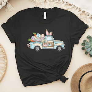 May include: Black t-shirt featuring a light blue pickup truck graphic carrying Easter eggs, carrots, and daisies. The truck has bunny ears on top and the word "Mama" on the side. The truck bed is filled with decorated eggs.