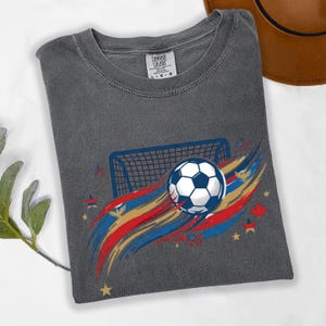 May include: A charcoal gray t-shirt featuring a graphic of a soccer ball and a goal net. The design includes red, blue, and gold brushstroke accents, along with small star and eagle icons. The inner neck label reads Comfort Colors size L-G-G.