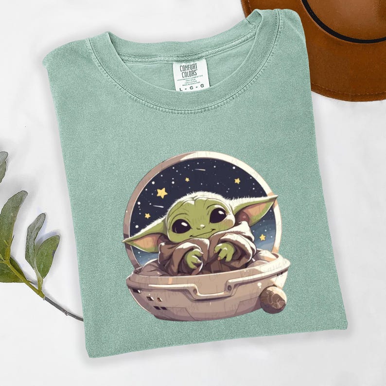 Puede incluir: Camiseta verde claro con una ilustraci&oacute;n de dibujos animados de un personaje alien&iacute;gena verde en una nave espacial sobre un fondo estrellado. La camiseta tiene cuello redondo y la etiqueta de la marca Comfort Colors.