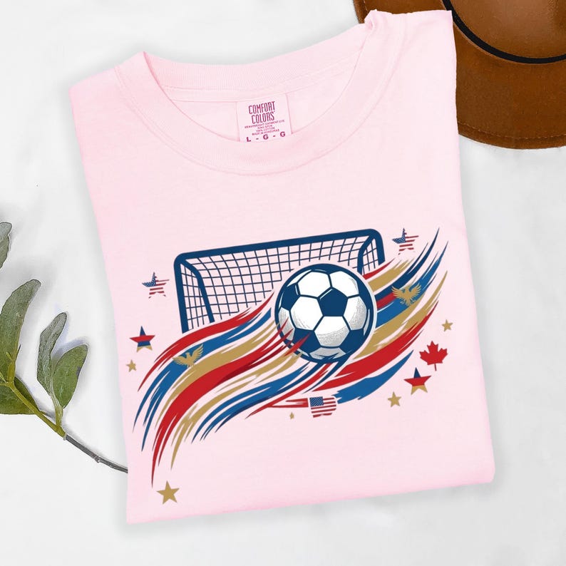 May include: A light pink t-shirt featuring a graphic of a soccer ball in front of a goal net. The design includes red, white, blue, and gold brushstroke accents, small stars, a maple leaf, and a small American flag. The neck label reads Comfort Colors.