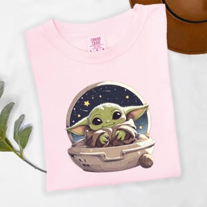 Puede incluir: Una camiseta rosa claro con una imagen de dibujos animados de un personaje alien&iacute;gena verde en una nave espacial contra un fondo estrellado. La camiseta tiene las palabras "Comfort Colors" y la talla "L-G" impresas.