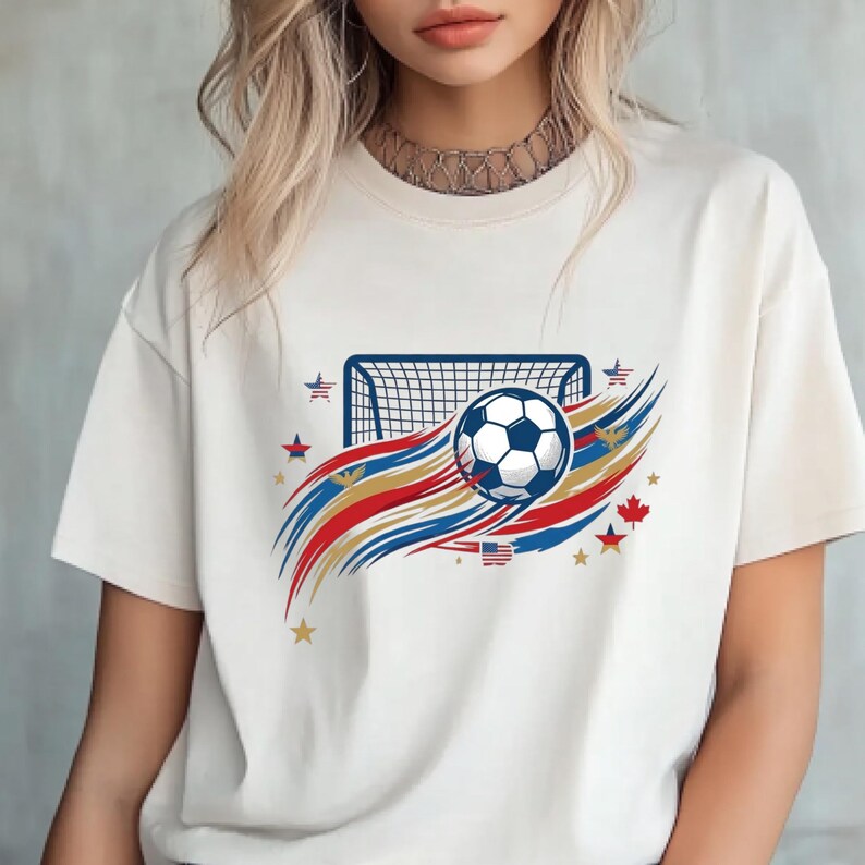 May include: A cream-colored short-sleeve t-shirt featuring a graphic of a soccer ball flying toward a goal net. The design includes red, white, and blue swooshes, small stars, a maple leaf, and a small American flag icon, creating a patriotic theme.